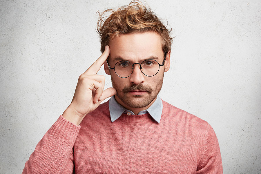 Photo of serious unshaven young male wears round spectacles, keeps finger on temple and looks strictly, tries to concentrate and make important speech, isolated over white concrete background